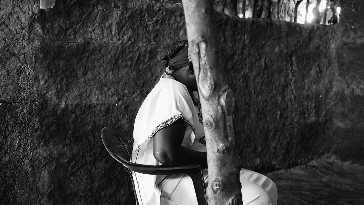 A black and white photo of a person sitting in a chair facing away from the camera with their faced obscured by a tree.