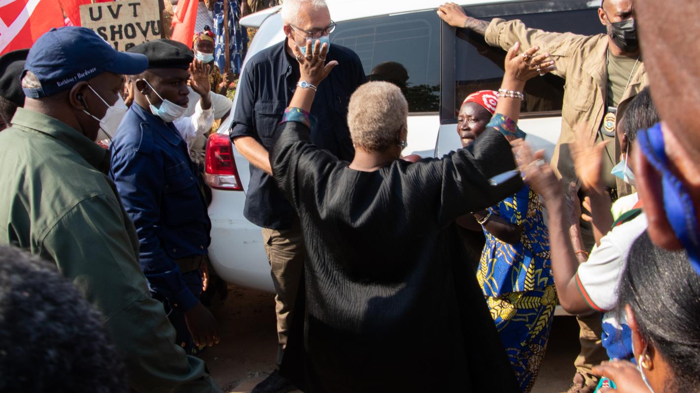 Person standing with raised arms in front of a white vehicle, surrounded by a group of people outdoors. Some individuals are wearing uniforms, and red flags are visible in the background.