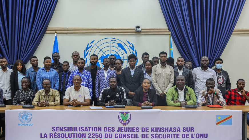 Un groupe de jeunes gens sont assis à un bureau et posent pour une photo devant l'emblème des Nations Unies.