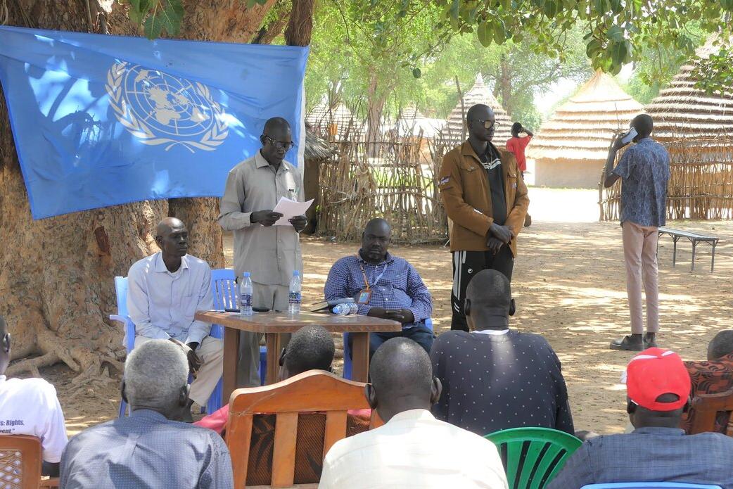 Youth leaders and other civil society activists Twic County have come together to spread the word of what previously feuding Twic and Ngok Dinka youth agreed on during a recent peace dialogue. Outdoor meeting under a large tree with a UN flag displayed. Several people are seated on chairs facing a small table where others are standing and speaking. Traditional huts are visible in the background.