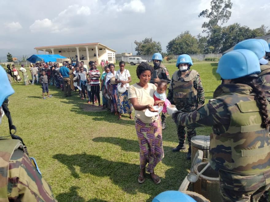 A line of people standing outdoors on grass, receiving aid from uniformed personnel wearing blue helmets marked “UN,” with a building and trees visible in the background under a partly cloudy sky.