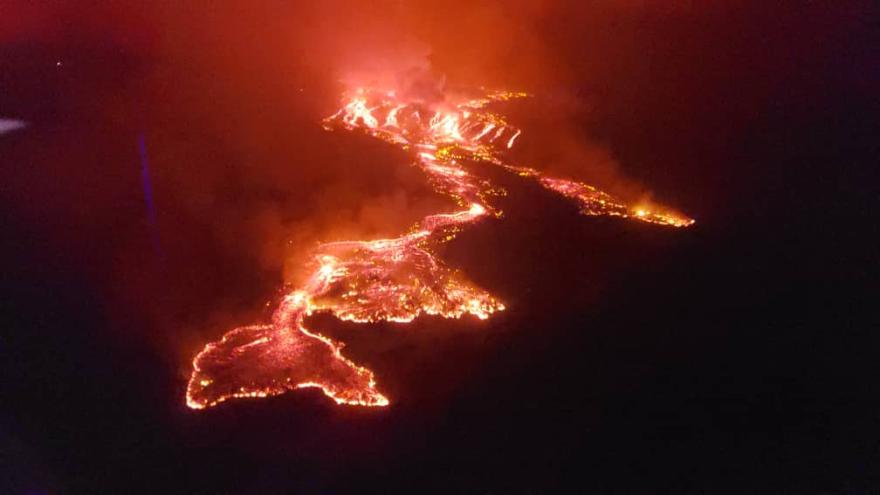 Aerial view of bright orange lava flows cutting through dark terrain at night, with glowing molten rock creating branching patterns and smoke rising from multiple points.