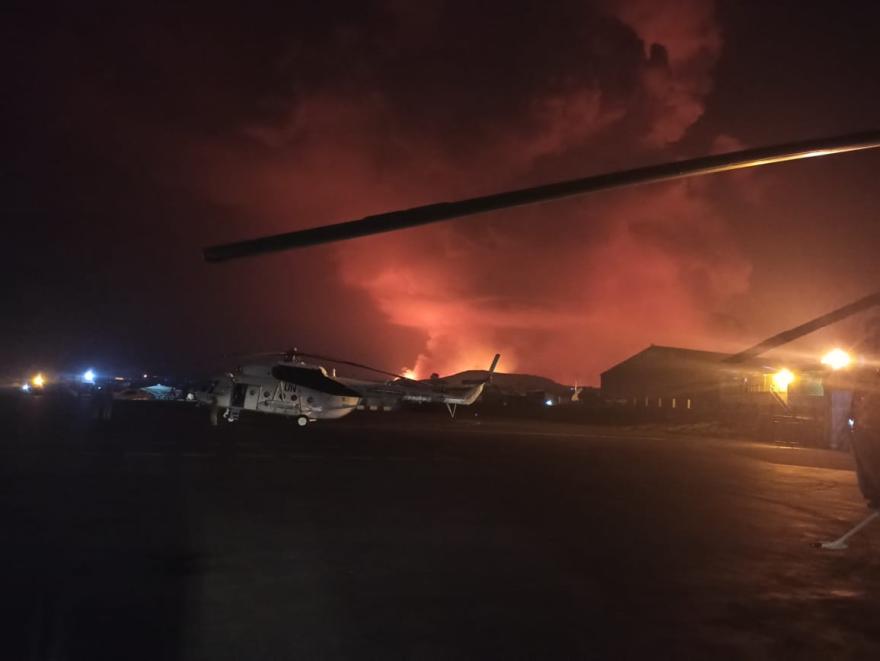 Helicopters parked on a dark airstrip at night with an intense orange glow in the sky from a distant fire or volcanic eruption, and buildings illuminated by artificial lights on the right.