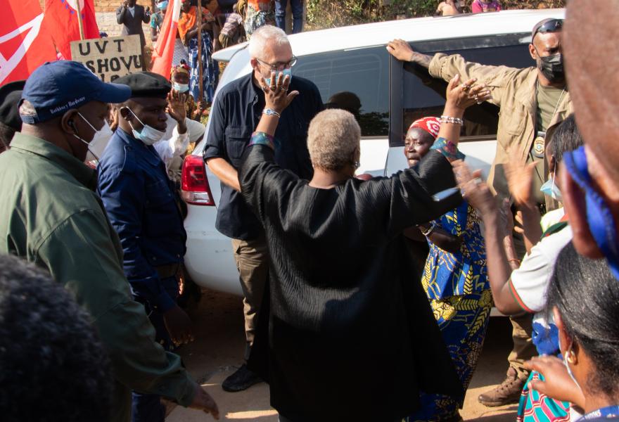 Person standing with raised arms in front of a white vehicle, surrounded by a group of people outdoors. Some individuals are wearing uniforms, and red flags are visible in the background.