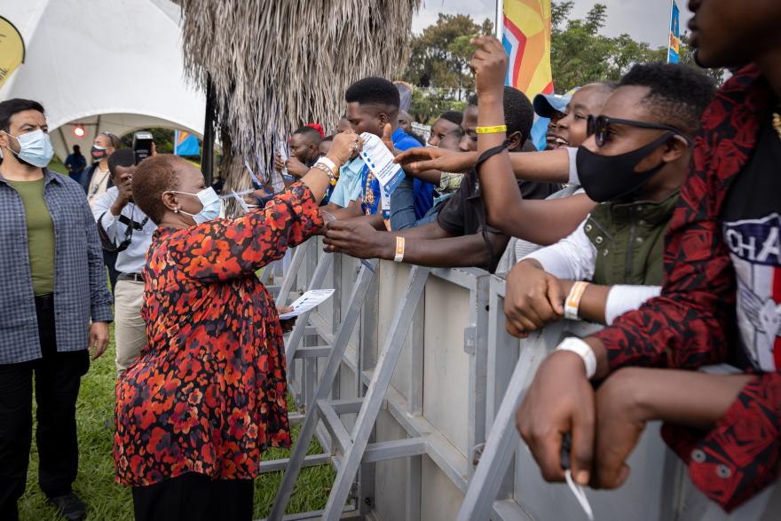 Person wearing a patterned top interacting with a crowd of people behind a metal barricade at an outdoor event, with tents and flags visible in the background.