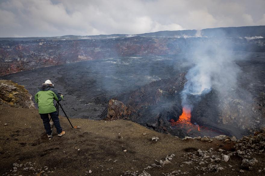 Person in protective gear standing near the edge of a volcanic crater, photographing an active vent emitting white smoke and glowing orange lava, with dark hardened lava covering the surrounding area.