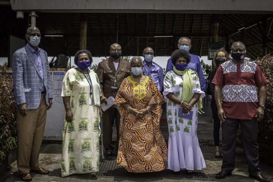 Group of eight adults standing in front of a building entrance with a thatched roof, dressed in colorful formal and traditional attire, posing for a photo outdoors.