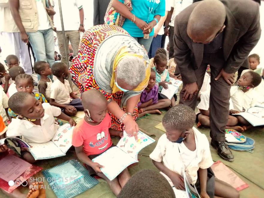 Children seated on the floor holding open books, while two adults lean down to engage with them. Other people stand in the background, and the setting appears to be an educational or community activity indoors.