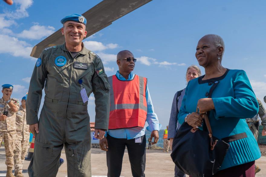Group of people standing on a paved surface outdoors, including uniformed personnel wearing blue helmets and others in civilian clothing, with part of a helicopter rotor visible above and clear blue sky in the background.