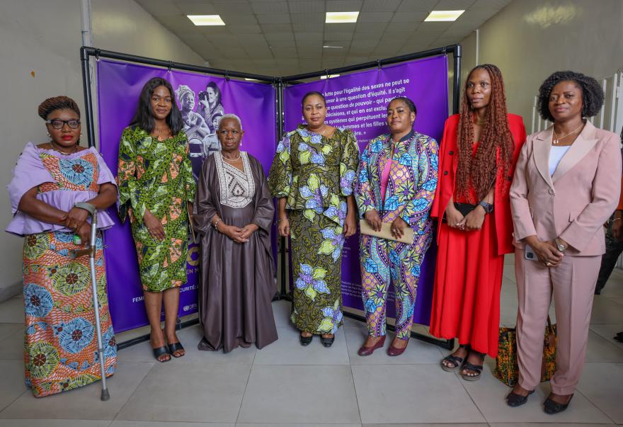 Seven adults standing indoors in front of a purple backdrop with text and images, dressed in colorful traditional and formal clothing, posing for a group photo.