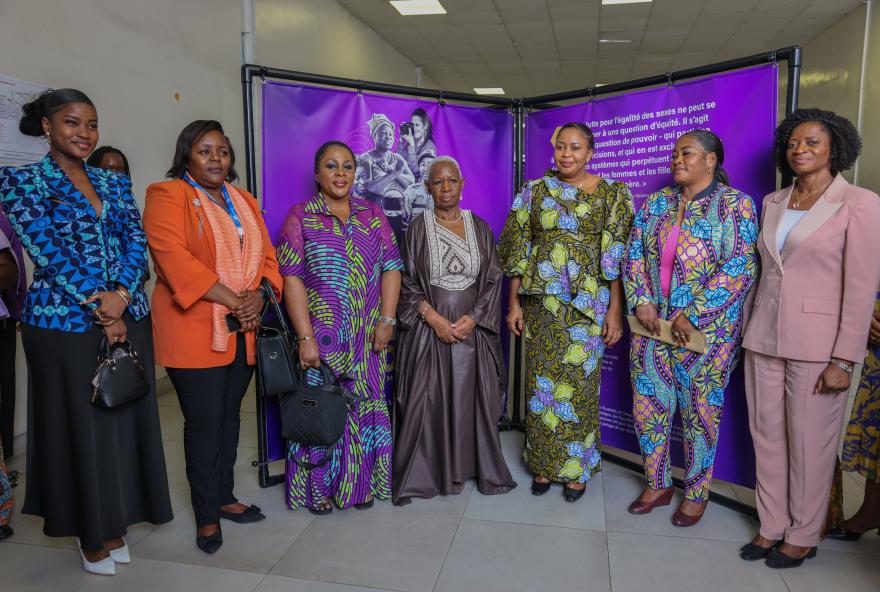 Group of seven people standing indoors in front of a purple backdrop with text and images, dressed in colorful traditional and formal clothing, posing for a photo